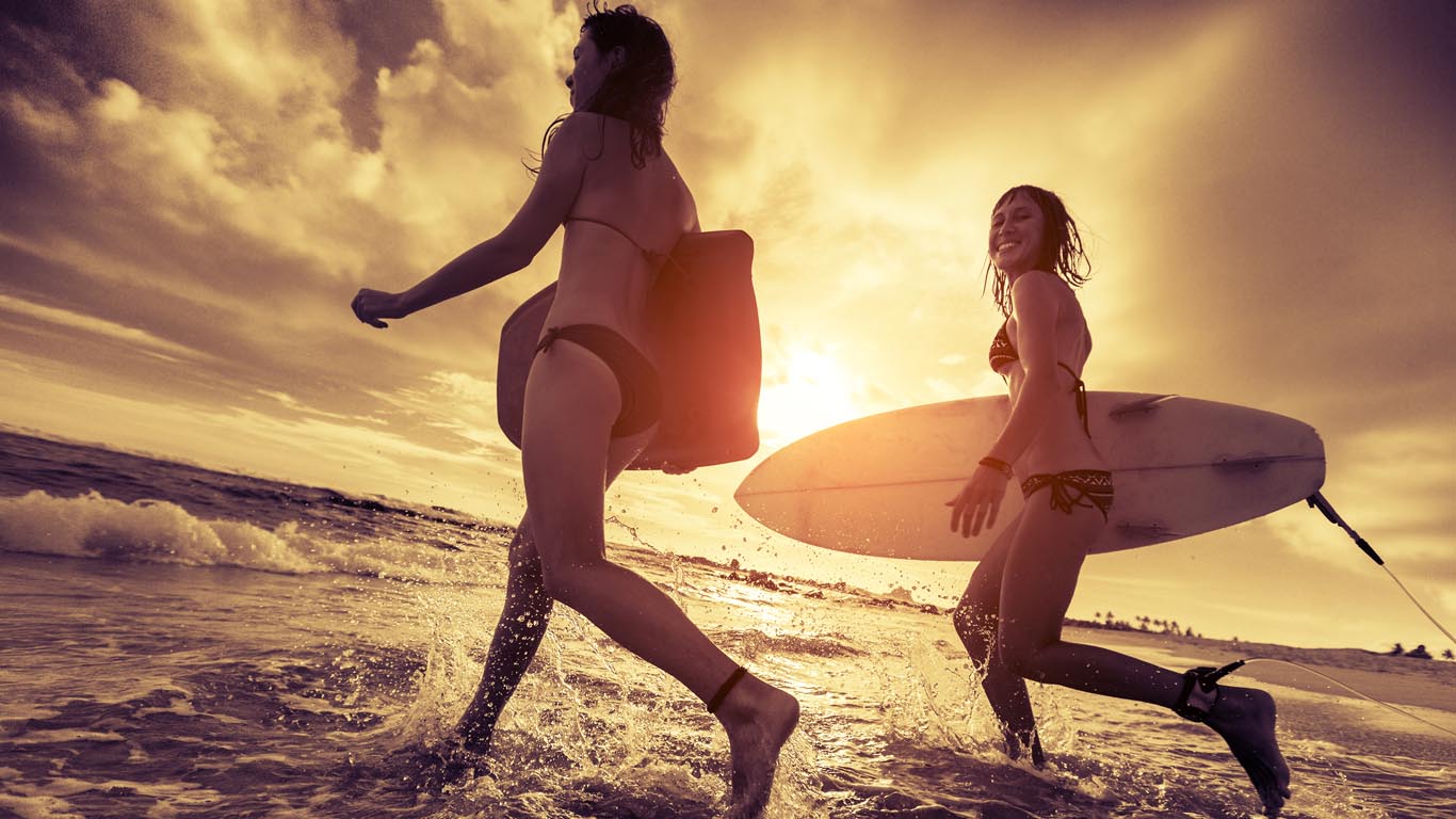 Two ladies surfers running with surfboards into the sea at sunset